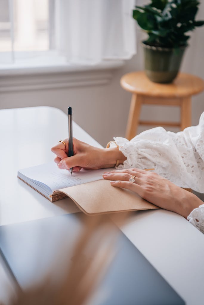 Close-up of a woman writing in a notebook at a bright home office desk.