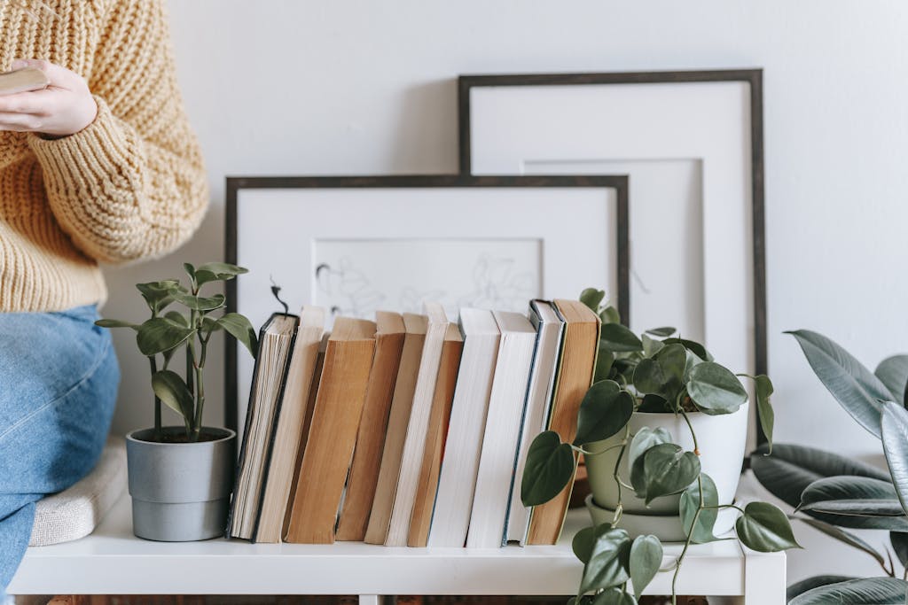 Crop female in casual clothes holding opened book and sitting on shelf with collection of books and plants and photo frames on white wall in light room