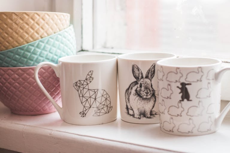 Elegant ceramic mugs with rabbit designs and colorful bowls on a windowsill, perfect for home decor inspiration.