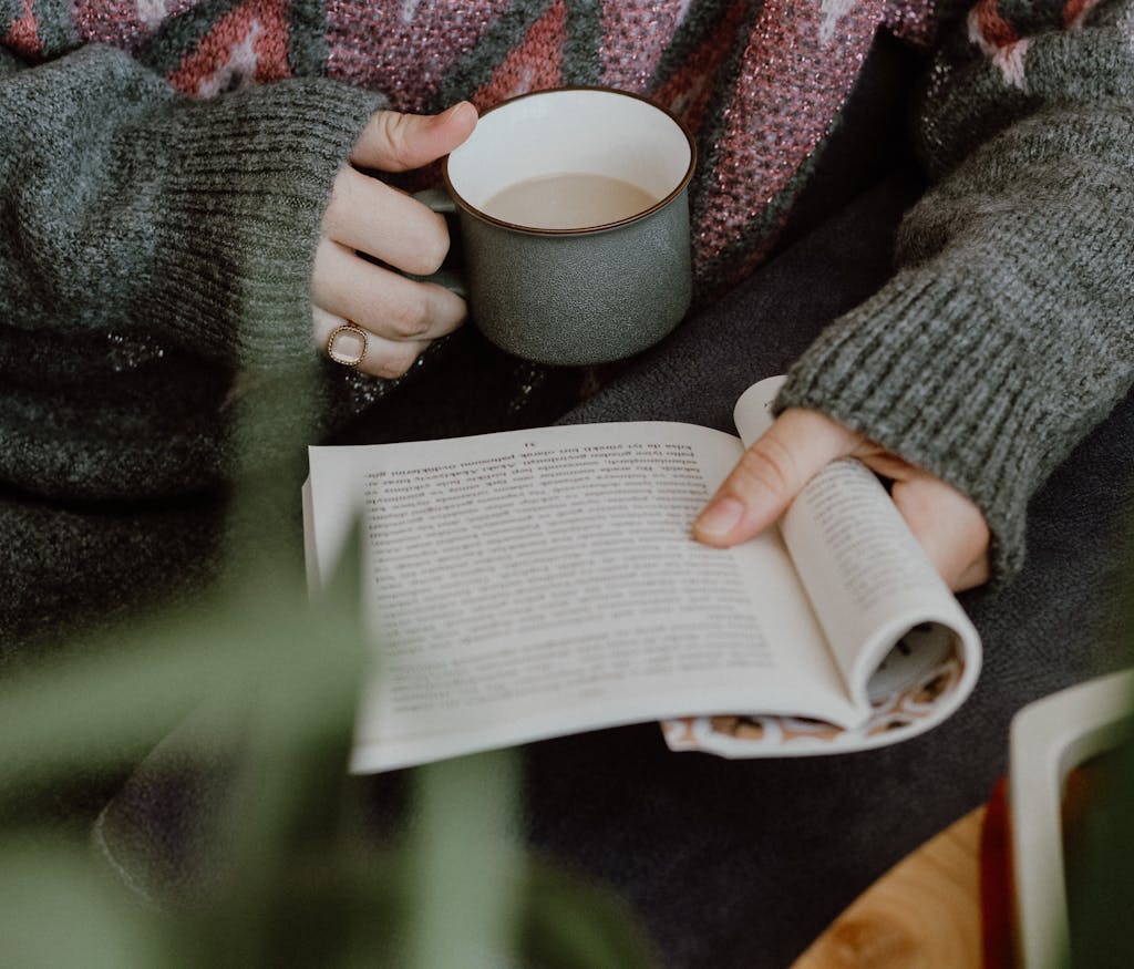 Person holding a coffee mug while reading a book indoors, exuding a warm and cozy atmosphere.